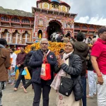 A happy senior couple standing with folded hands in front of the colourful Badrinath Temple during their Chardham Helicopter Package.