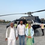Three happy passengers standing together on the tarmac right next to a premium dark blue helicopter during their 4 Dham yatra by Helicopter 2026.